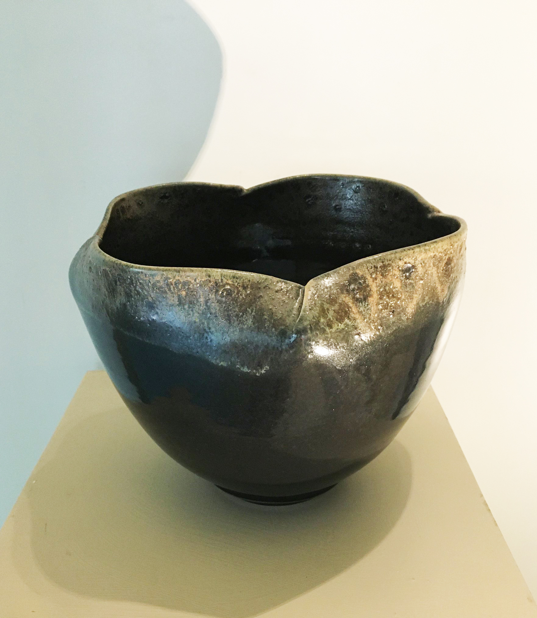 A ceramic bowl with areas on the lip of the bowl that have been pinched. The glaze is dark on the bottom, but the rim has a mottled, lighter golden color.