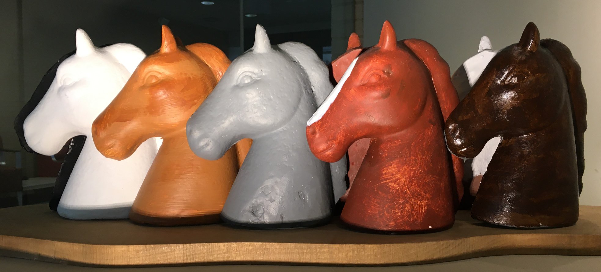This sculpture has an organic-shaped, gold-painted wood base that holds at least 8 horse heads. They are cast the same, but painted differently. From left to right: a dark head in the background, a white, orange, gray, red with a white blaze, and brown horse heads are near the viewer with an additional red and white horse are mostly obscured in the background. 