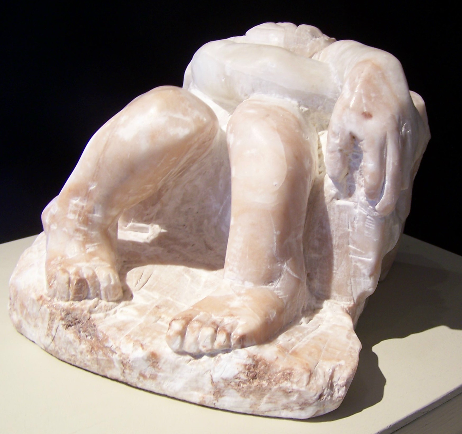 This is a sculpture of a human carved from marble. The figure is sitting down, almost submerged in the rock. The knees and feet are separately carved, emerging from the stone. The head is looking down, resting in the arms while the arms are crossed and rest above the knees to their right on the stone. The right hand is carved and almost grips the rock. The feet are angled in towards one another. The toes on the left foot may have been broken off. The stone is roughly hewn and the overall feeling is of the figure almost sinking into the stone, of hiding or of rest. 