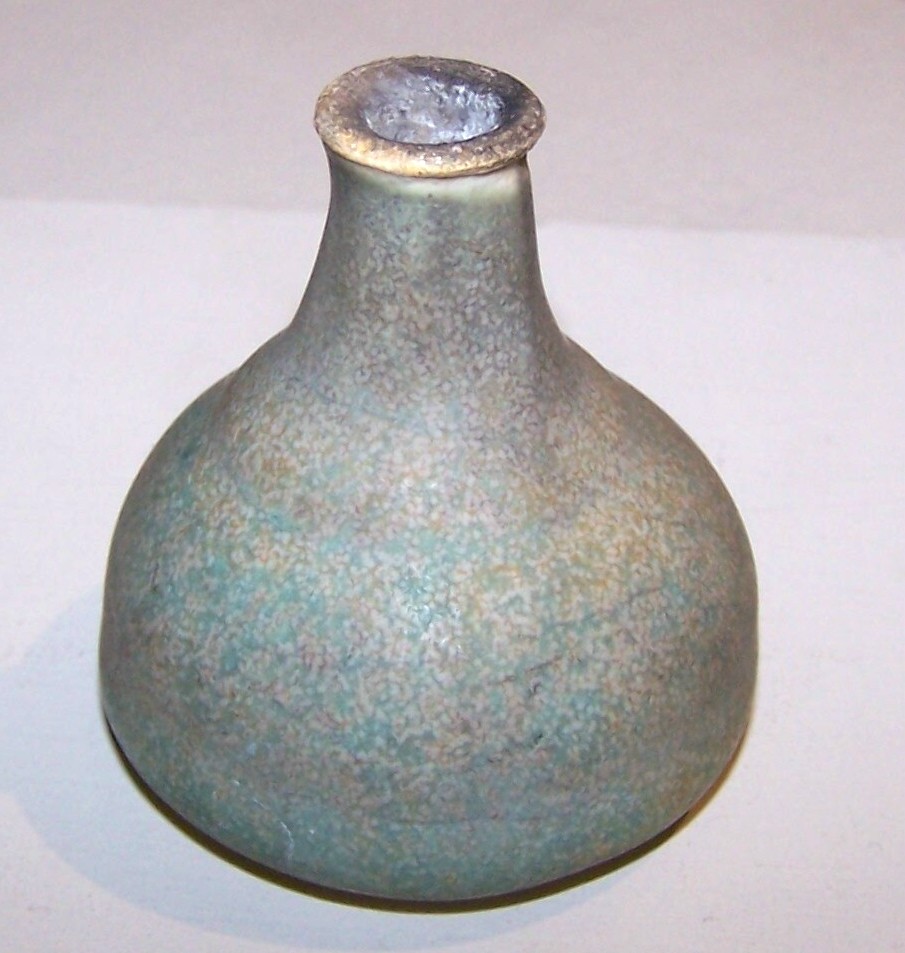 This is a pot or vase that is larger on the base, with a taller, narrow neck. It has a green and brown matte finish. The top of the vase is slightly wider than the neck and the interior has an ashy appearance. This may have been a raku-fired piece.