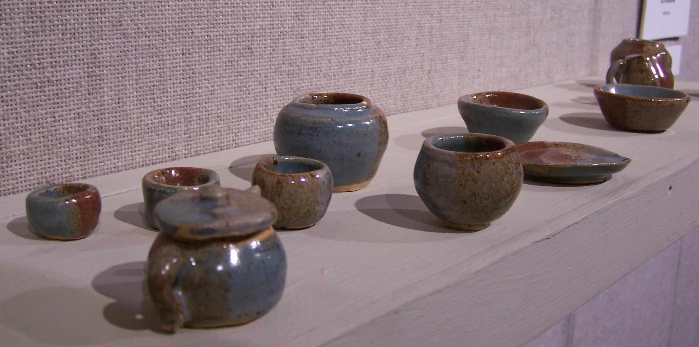This photograph contains a shelf holding eleven ceramic forms. There is a teapot, four differently sized cups, a squat vase, a plate, two bowls, a pitcher, and another vase. The glaze consists of blue, red and brown tones. The forms are imperfectly made, a little wobbly, but a very cohesive set. 