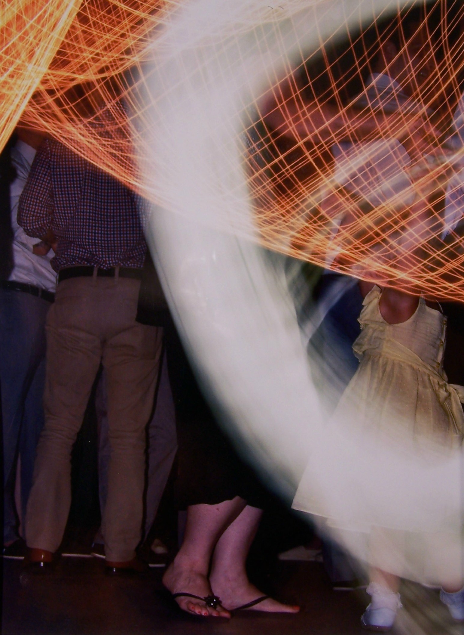 This color photograph is taken at some type of dance, possibly a wedding. There are multiple people dressed in business casual in the background that are mostly obscured from view by a large white semi transparent swoosh of white that moves from the top right and curves down to the bottom right of the work. The top third of the piece also contains multiple areas of orange lines in a grid-like pattern, similar to a 3D mathematical plot. There is a woman twirling a small girl wearing a light-colored dress.