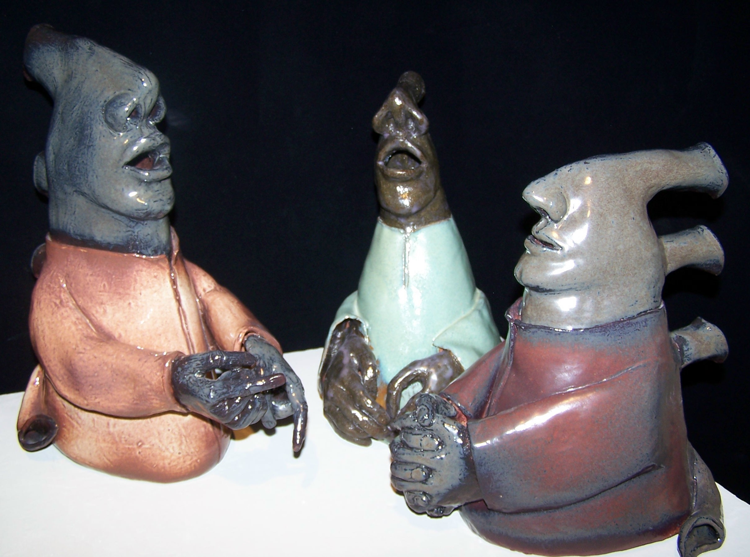 This image contains three ceramic sculptures of humanoid figures with their mouths open to varying degrees. Looking straight on, the pieces have a triangular structure to them where the base is wider than the top. The figures tops begin with the nose that reaches to the back of the form. They have mouths and chins leading to a thick neck but no eyes, ears, or forehead. The figures end at the waist and each have arms that are reaching/clasped in front of them. They are all facing a similar point in the frame, slightly angled towards one another. On the back of each are four tubes. Three reach back from the nose, the back of the neck, and the upper back, which the fourth curves to the ground. These could be possibly musical instrument pipes or simply a design element. The figure on the left has blue-gray skin with an orange shirt. The middle figure has brown skin with a teal shirt. The right figure has gray skin with a blue-red shirt.