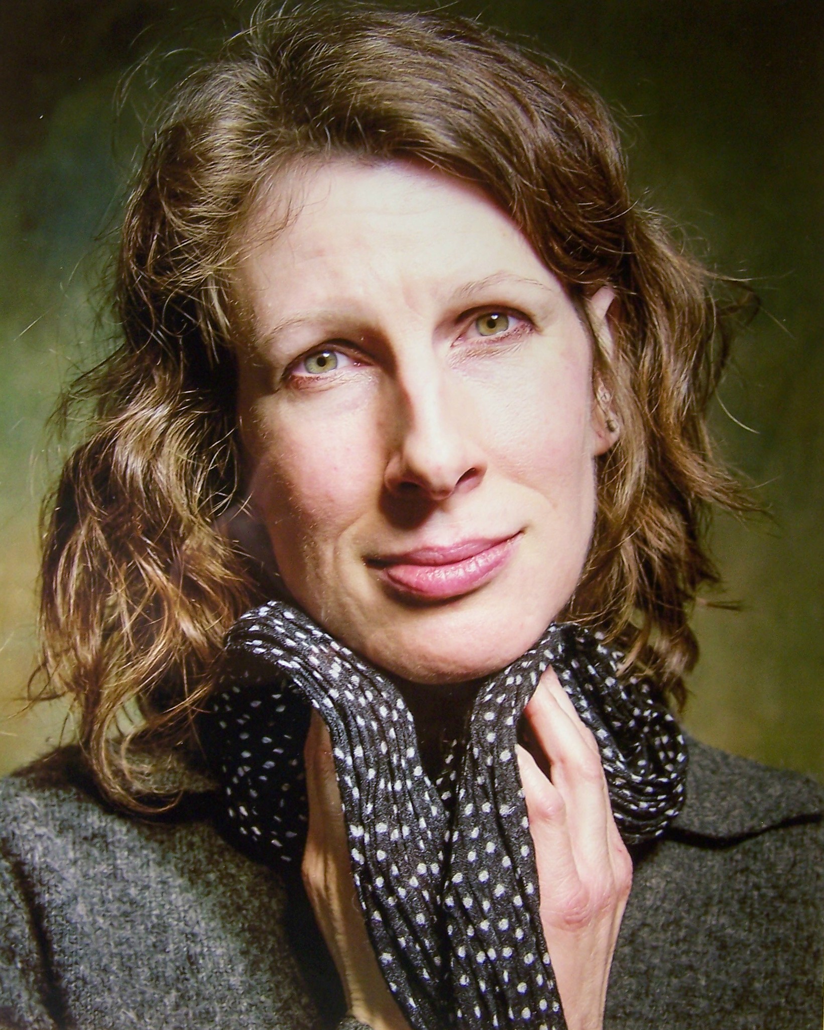 A portrait photograph of a woman holding a polka-dot scarf up to her chin. She has shoulder-length wavy brown hair and a soft expression that stares at the viewer. She has a tall oval face and wears a gray wool-like sweater. A yellow-green background is brightest by her shoulders and fades to darker hues towards the top of the image. It echoes the colors in her eyes.