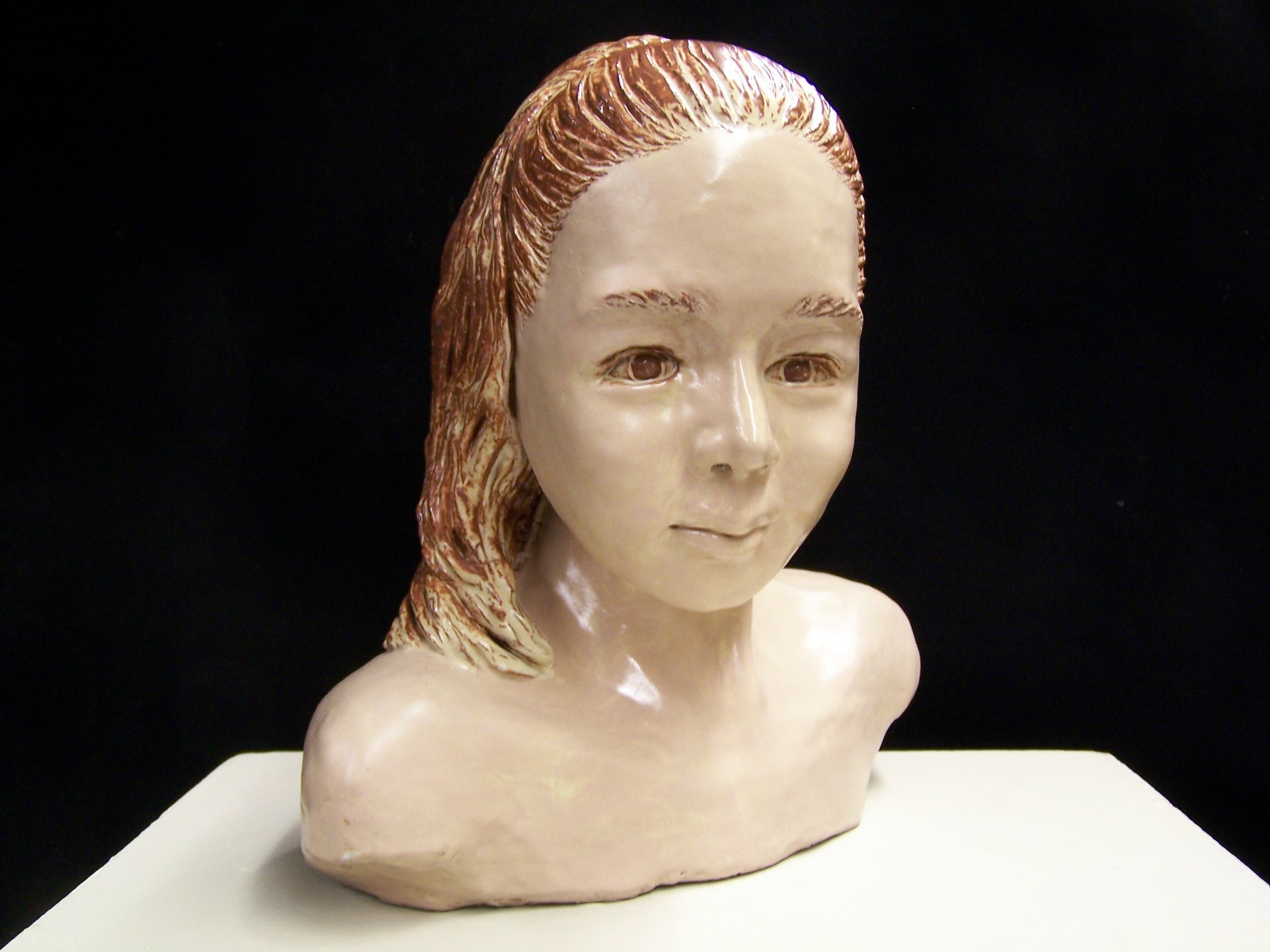 This pieces is a glazed ceramic bust of a young woman. Shown from the armpits up, she is two-toned with creamy skin and dark red-brown accents in her eyes, eyebrows, and hair. Her hair is up in a ponytail, which rests on her left shoulder. She has a bit of an impish look on her face. 