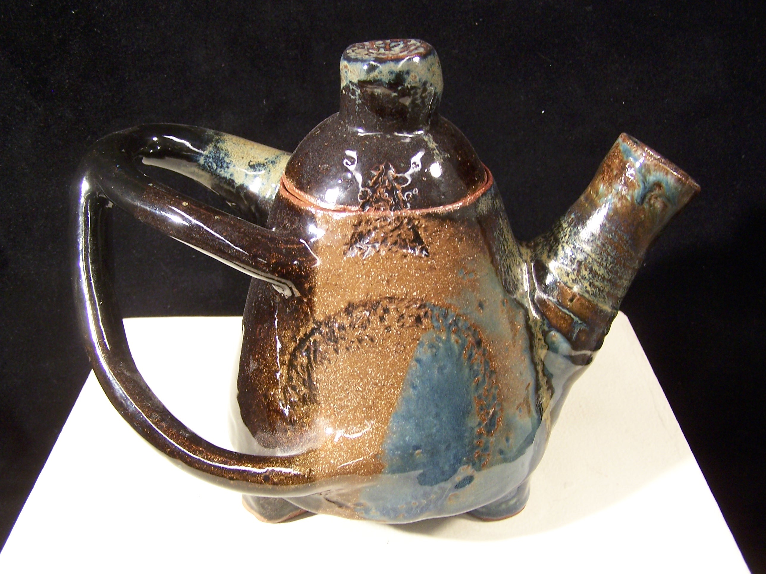A glazed ceramic teapot. It is roughly egg shaped with two visible short feet attached to the bottom. The sturdy spout appears to be wheel-thrown and is angled upward from near the base. The lid has a sizeable knob. The handle is uniquely shaped. The top is a u-shape, connected to the pot relatively near the lid towards the back of the piece. One arm comes off the middle and swoops to connect to the base of the pot. The glaze is dark brown, light brown, blue and a light ochre. There are some patterns on the side of the pot: a texturized circle and a triangle with an arched base that connects the pot to the lid. The top of the knob is also texturized.