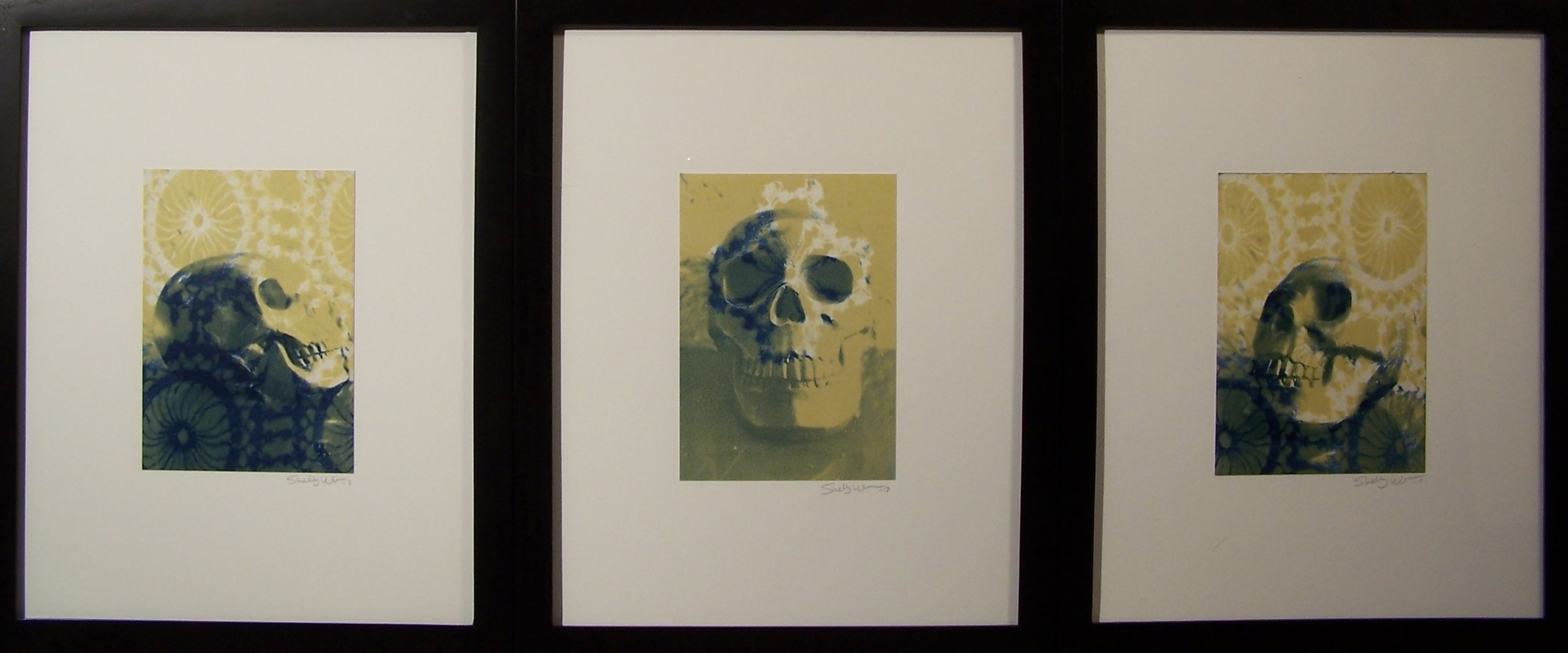 A triptych of three photographs of skulls. The far left image has a skull profile looking up and to the right with an overlay of pattern featuring a circles with lines moving to the center of them and patterning outside of them. The center one has the skull facing the viewer with lower contrast than the other two. The one on the right has a three quarter view of the skull looking up and to the left with the same overlay patterning as the far left. The colors on all three of them are white, yellow, and a dark blue. 