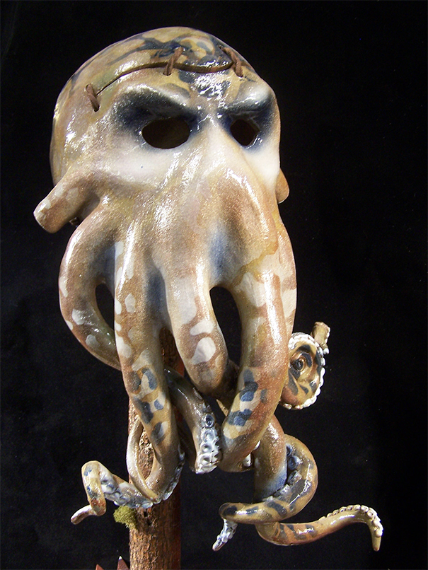 A ceramic representation of a kraken-like figure. The eyes are hollow with a furrowed brow above them, giving an angry or stern appearance. The bottom of the piece has five or more tentacles that curl around themselves. The forehead has a slit cut in it and is bound by leather or a thick rope