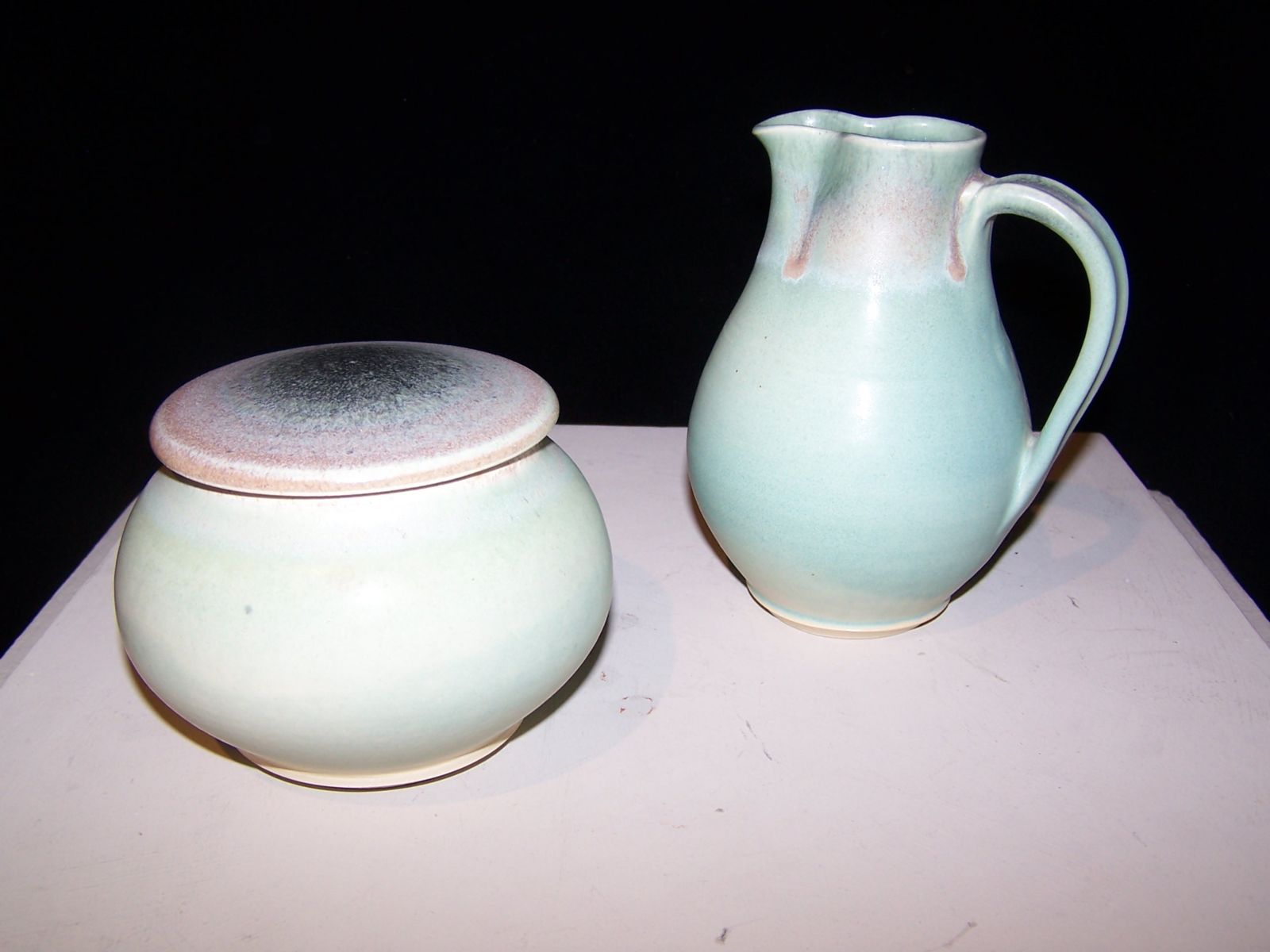 A ceramic sugar bowl with lid and a creamer pitcher. The Sugar bowl is round with a small foot and a low-profile lid without a handle. The creamer pitcher has a wide belly, angled foot, and narrows towards the top with a spout and handle. The glaze is a matte green that appears to break red and black towards the top of the pieces. The pieces appear to be wheel-thrown.