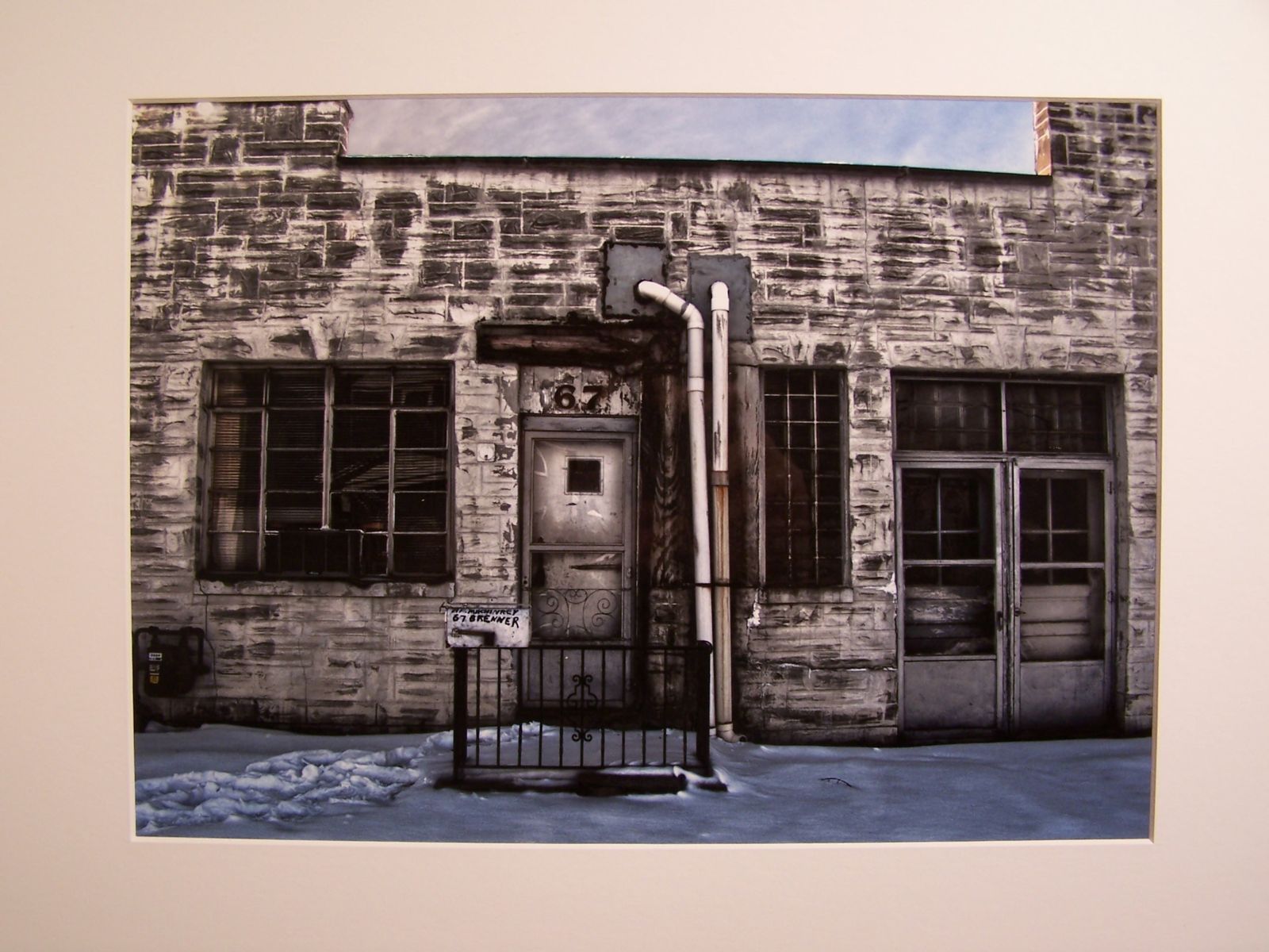 A photograph of an old stone building. There is a door with a small wrought iron fence around the stoop just to the left of center with number 67 above it. The fence holds a mailbox that has some undistinguishable writing above “67 Brenner.” A line of footprints in the snow lead to the door from the left. There is a large window to its left with multiple panes and a vertical paned window to its right. There are double doors further to the right. To the immediate right of the central door are two PVC pipes going from above the door to the ground. The building’s façade continues up on the left and right of the photograph, but reveals a long, rectangular sliver of the sky in the middle. The photograph is full color, but the colors are all so muted, it reads almost black and white from first glance. Closer inspection reveals the snow and sky to be cool tones and the building to be warm and a gas meter on the far left has a yellow tag on it.