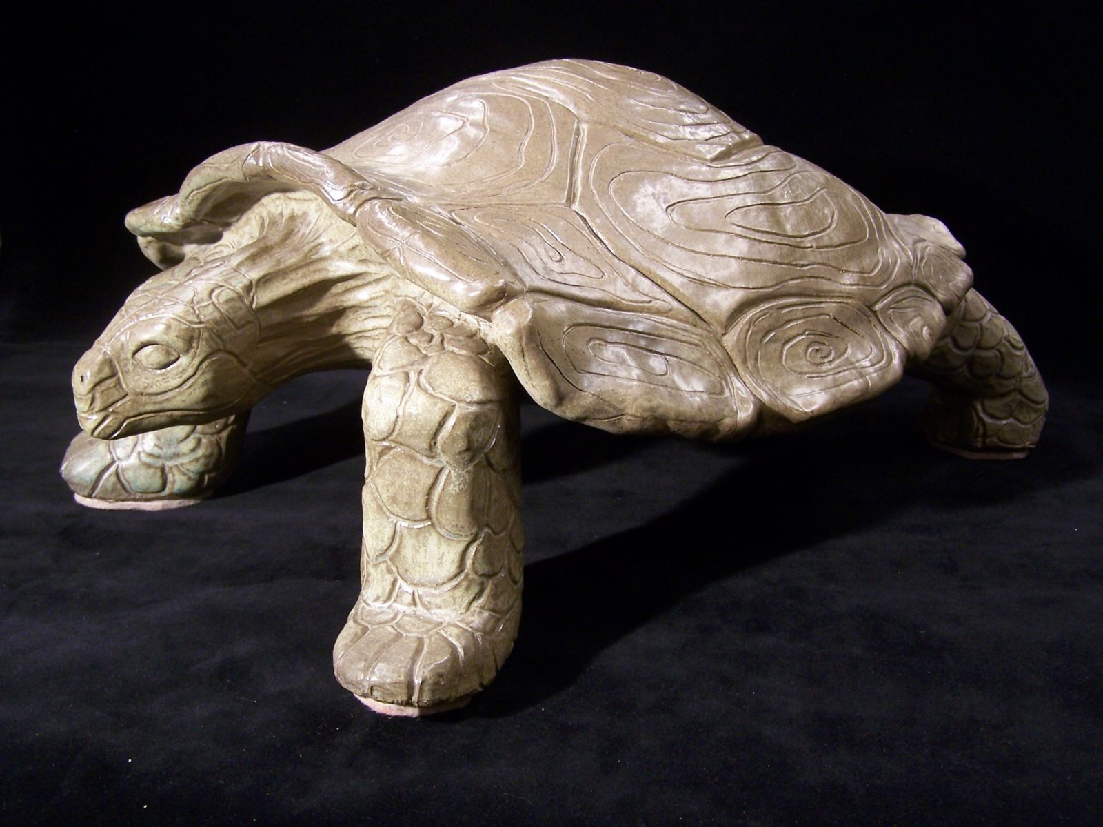 This sculpture of a stylized tortoise appears to be ceramic. It has a sheen and what looks to be subtle variations of a light brown glaze over the entire piece. The shell has swirls carved into its sections, the legs have scales carved into them and the head has a similar scale pattern conforming to the sections of the face. The tortoise’s head is stretching forward and slightly down. It’s a very organic, fairly realistic depiction of the animal.