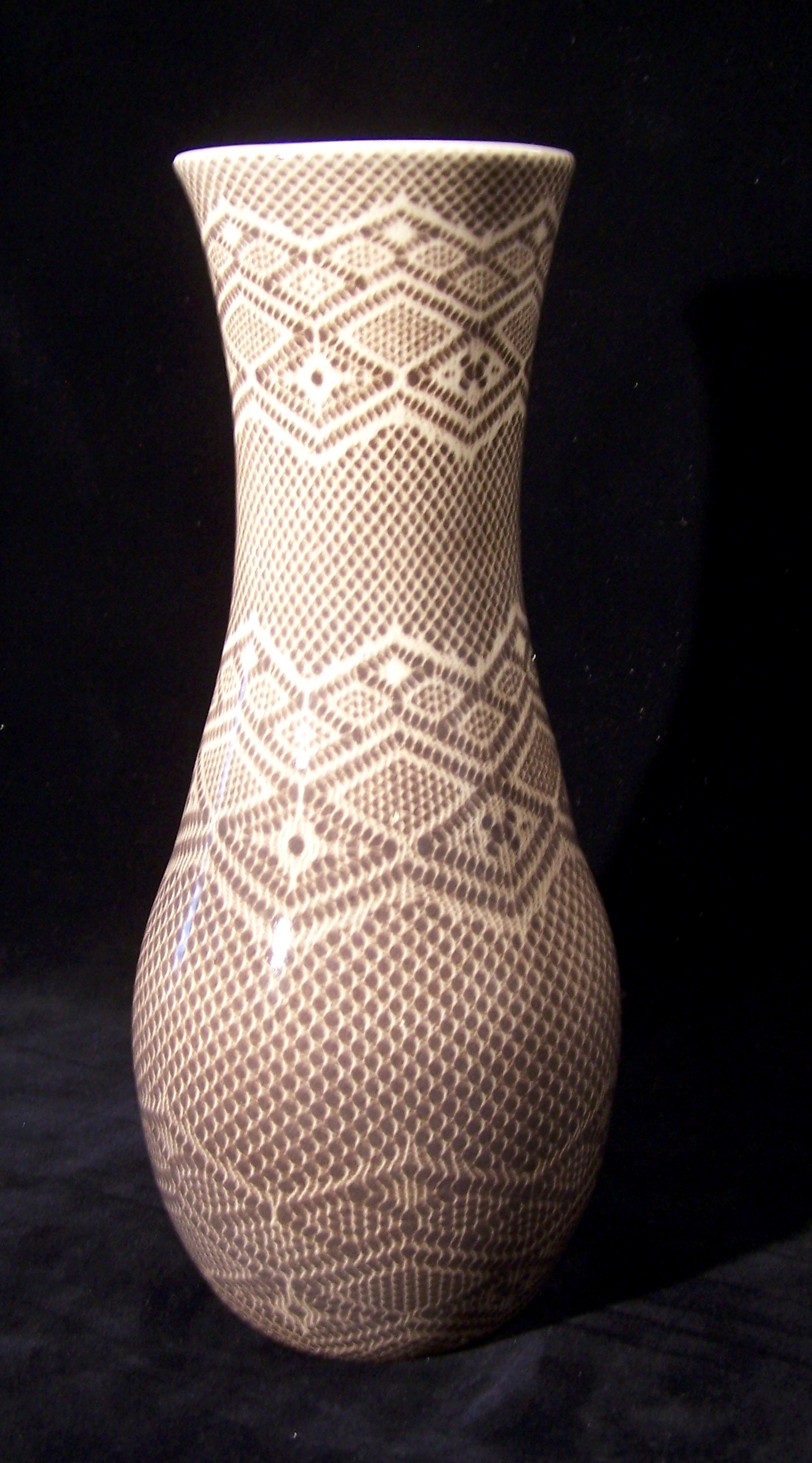 A tall, thin vase with an intricate pattern carved or impressed into the entire surface that replicates a lace or knit fabric. Elongated diamonds of various sizes are a repeated motif in three bands around the top, middle, and bottom of the piece. It appears to be a white base with brown glaze inside the impressions.