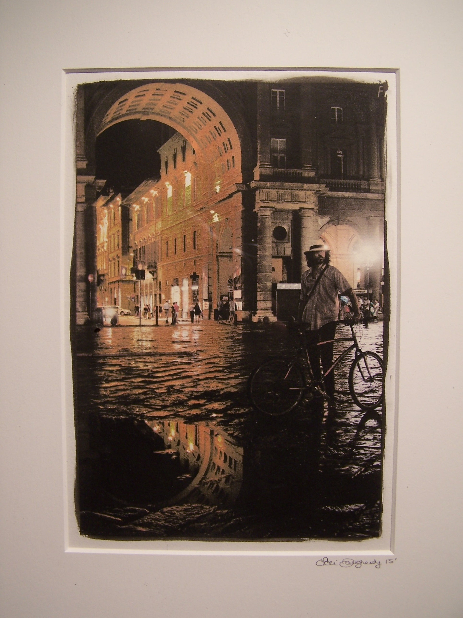 This black and white photograph features the Piazza della Republica in Florence, Italy after the rain. Its large architectural arch is on the left side of the frame with buildings on the right and dark, wet cobblestones in the foreground. A puddle in the foreground is reflecting the arch. A man with a fedora and bicycle looks off frame to the right. The arch and other moments of the photograph are colored with salmon and mustard green hues with other parts remaining black and white.