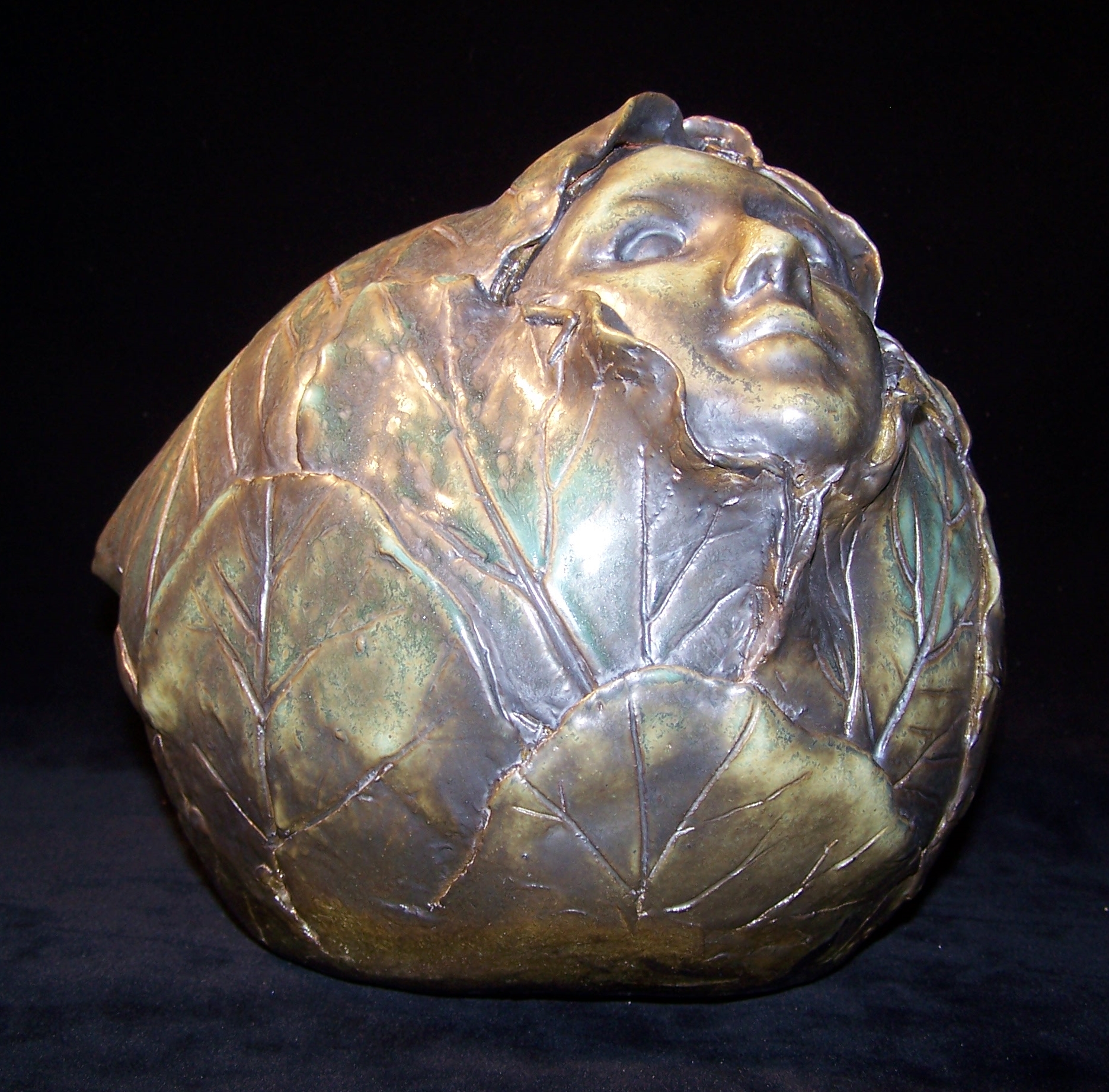 A sculpture of a head of lettuce. It has an iridescent finish of gold, green and brown. It appears to be made out of clay. Out of the top side a face is emerging. It is smoothly rendered and is framed by the leaves of the lettuce.