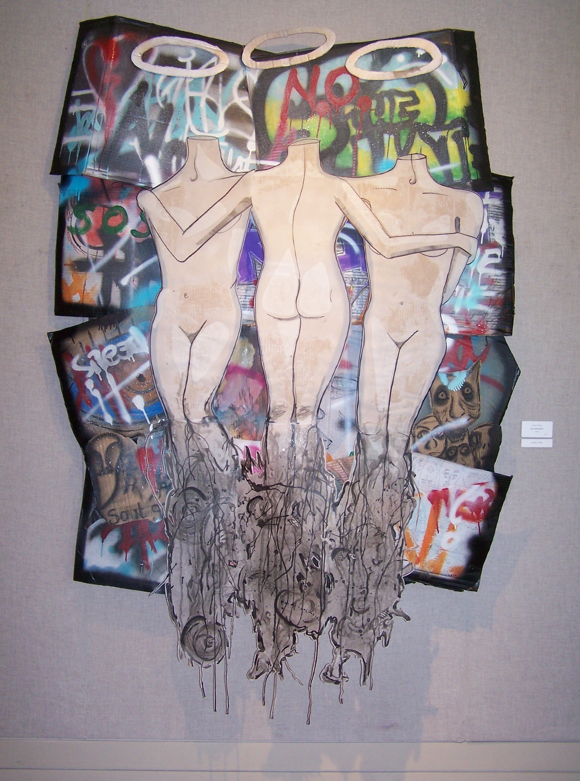 This collage is a 2D freeformed collage. The background is a handful of collaged cardboard rectangles that have been spraypainted in graffiti in black, green, yellow, blue, red and orange. The cardboard in some places is left showing. On top of that are three headless female forms. The ones on the right and left are facing the viewer and the middle is facing away with her arms on the shoulders of the others. At the knees their bodies dissolve into unrecognizable melting/dripping scribbles that extend beyond the cardboard backing. Three halos float above where their heads would be. 