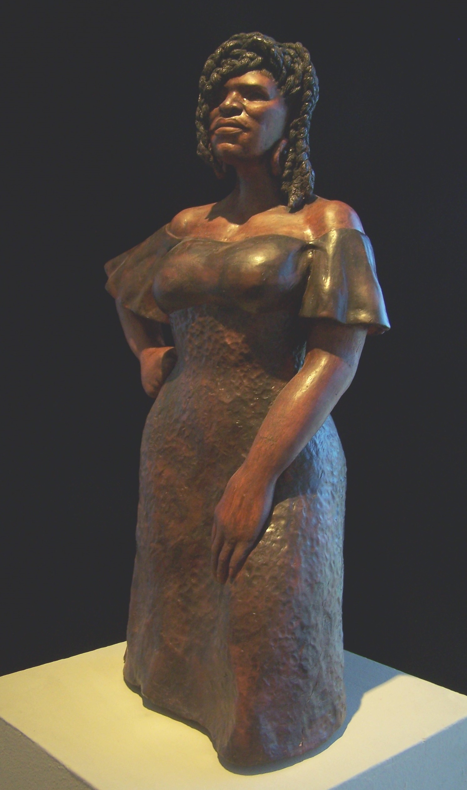 This sculpture, probably clay, is of a full figured black woman portrayed from the mid-leg up. She is wearing an off-the-shoulders dress and her weight is resting on her right leg. Her right arm is at her hip and her left arm is resting on her leg. She is looking slightly up and has a proud aura to her. Her hair is in thick braids that rest on her shoulders.