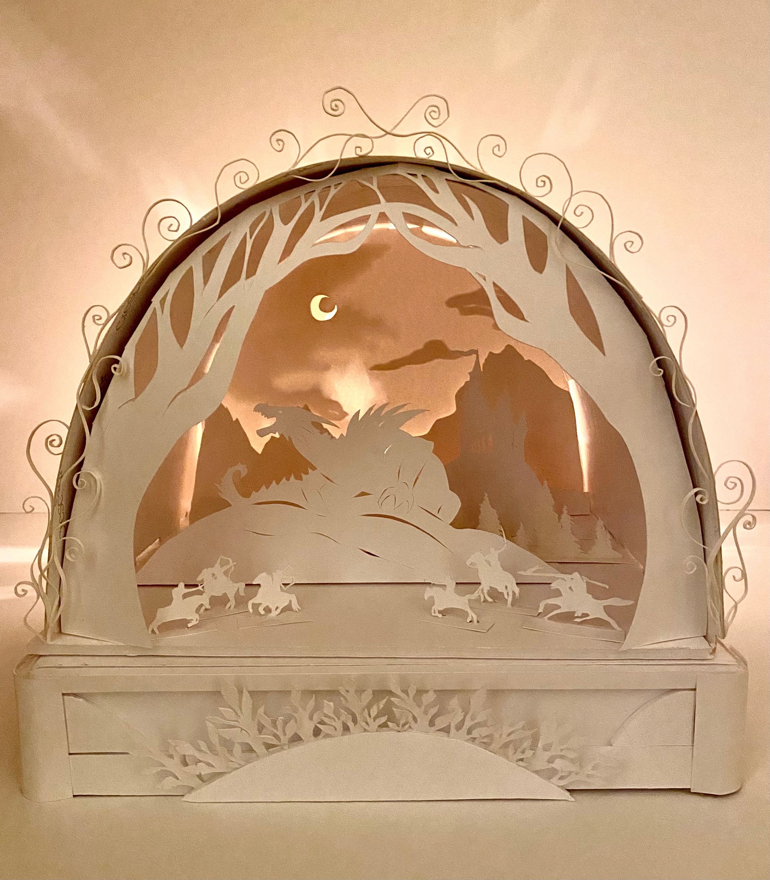 This 3D paper diorama has an embellished rectangle platform with a scene depicted on top of it enclosed in arched paper. The foreground has 2D trees growing along the front of the arch and quilled paper breaks the bounds of the arch. Inside is a mountainous night scene where a large 2D dragon is roaring over small figures riding horseback with weapons extended. The entire piece is dramatically backlit and the different depths of cut paper reveal a bright crescent moon, dark clouds, and mountains in the background.