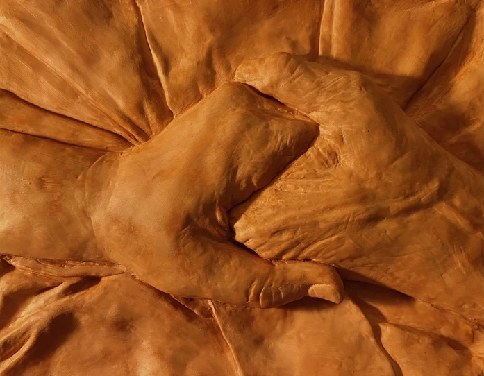 A red clay relief showing two right hands clasping each other. The right is aged while the left is a younger hand. The hands rest on a cloth.