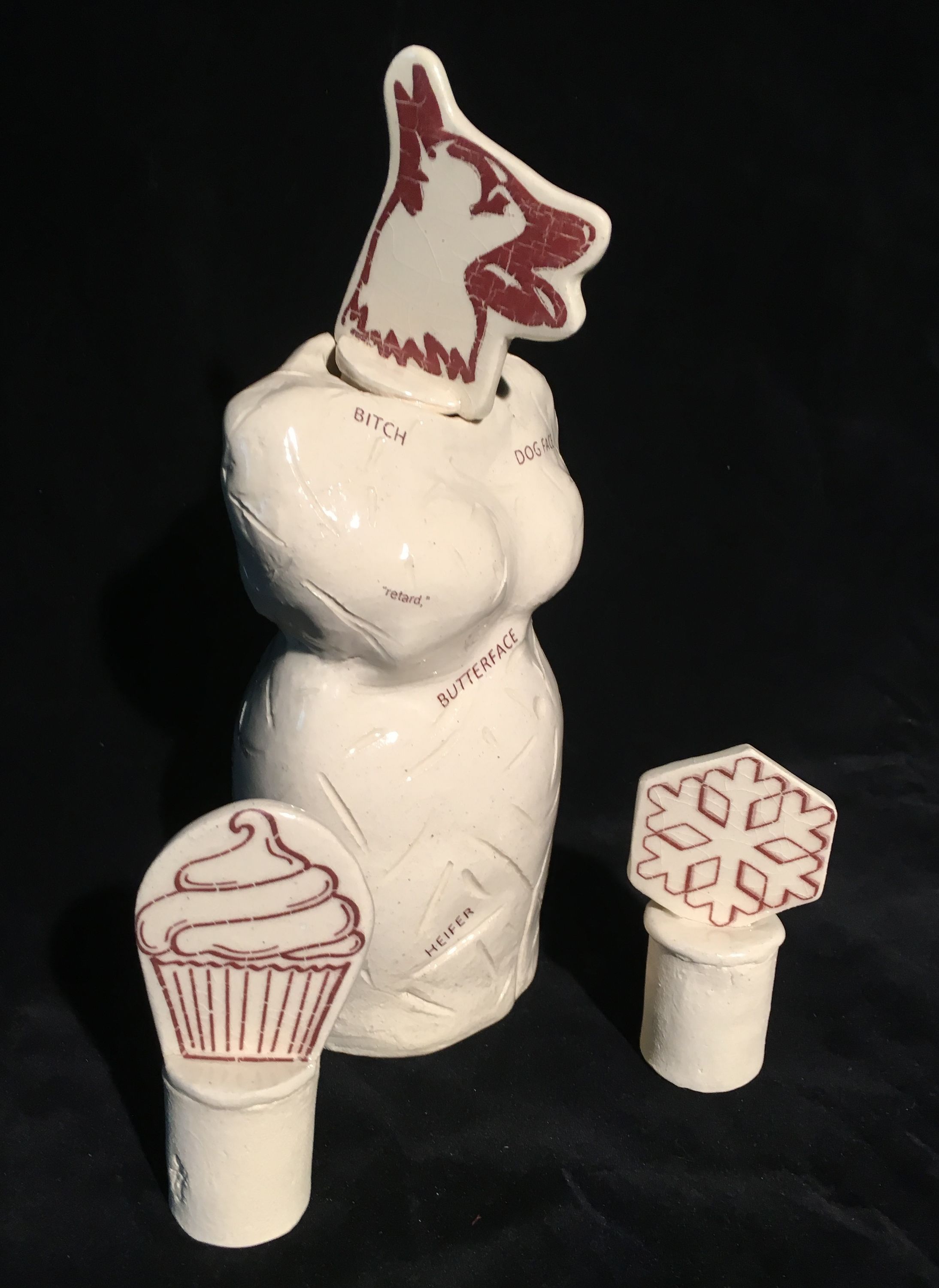 This ceramic sculpture is an abstracted woman’s form from neck to thighs sans arms. The form is marked with slashes and small words are printed on the surface in various sizes. Words include “BITCH, DOG FACE, retard, BUTTERFACE, AND HEIFER.” The head is interchangeable with 2d- inserts. The one shown is a German shepherd-like dog, but a snowflake and cupcake are additional options shown. 