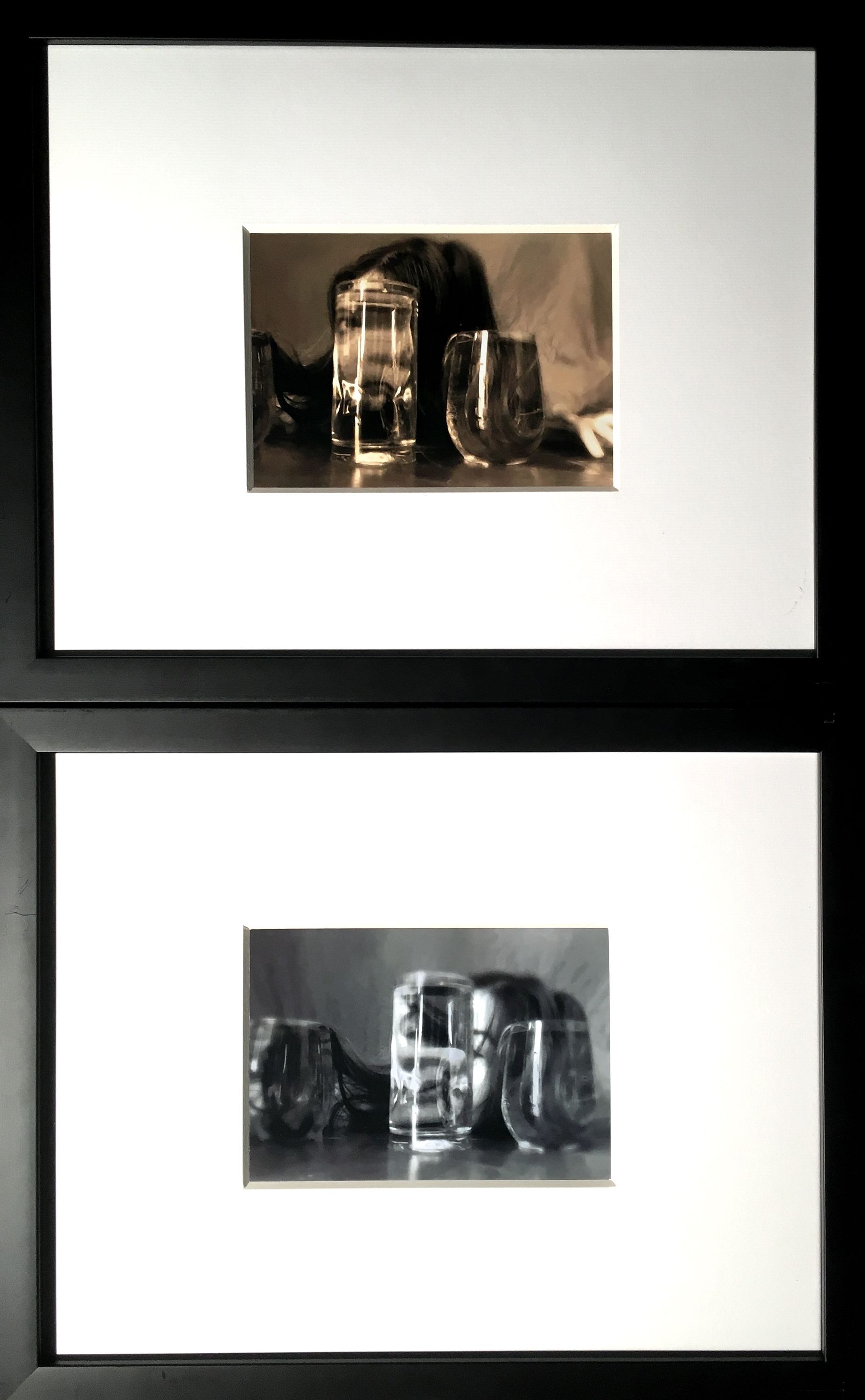 This vertical photo diptych has a still life of clear glasses of clear liquid in front of a person with long dark hair. In the top piece, the face, looking at the viewer, is distorted in the central glass and the hair is in the other two glasses. It is sepia toned. The second image is black and white. The head is tipped back a bit more and the reflection in the liquid is facing the figure who is mostly obscured by the glasses. 