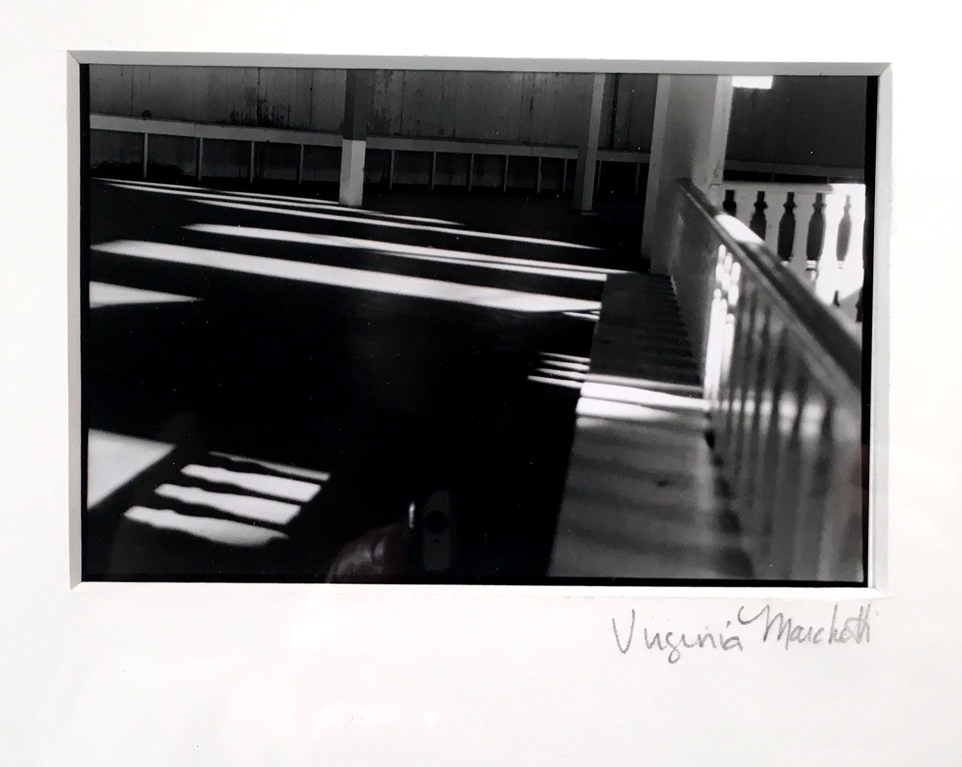 A black and white photograph of an interior space that’s a bit run down. Overall the composition is very geometric. There is a railing on the right-hand side and the rest of the space is open with a lot of blank floor moving to a back wall. The framing is at an angle to the horizon, so there are a lot of angles made by the railing, support posts, and back fence against the far wall. There is a light source on the right casting mostly shadows onto the space, but long rectangles of light pattern the floor and light passing though the railing continues the patterned repetition closer to the viewer. 