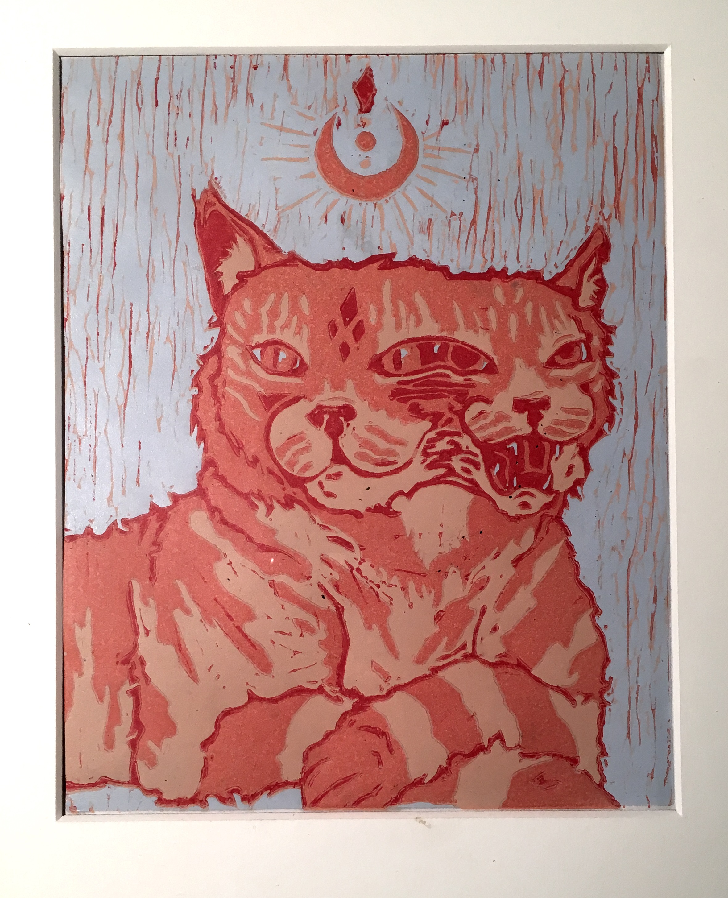 This print of a cat has a light blue background. The cat is laying down, shown from the torso up and is printed in three shades of salmon red. Chatter marks of the print block are visible throughout the print. The cat has two faces. One is relaxed and looking towards the left and the other is angry, mouth open and looking to the right. The faces are connected as if the cat was in motion or dividing itself into two. Above the cat is a crescent moon shape with rays coming off of it with a circle in the center and a diamond at the top. The cat’s head on the left has four diamonds in a diamond shape on its forehead.