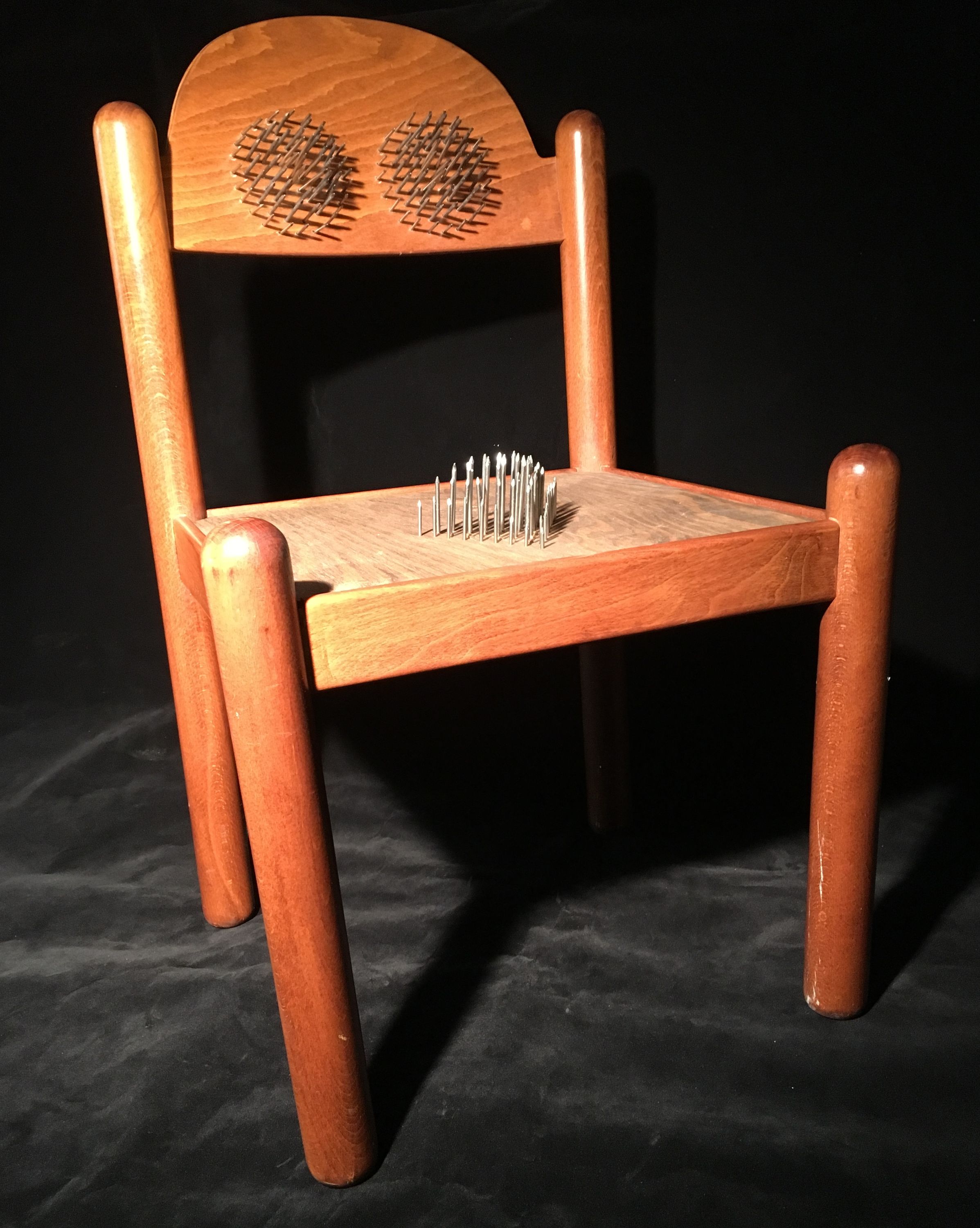 This uninviting sculpture is a simple wooden chair that has nails coming out of the back of the chair in two circles and one triangle on the seat.