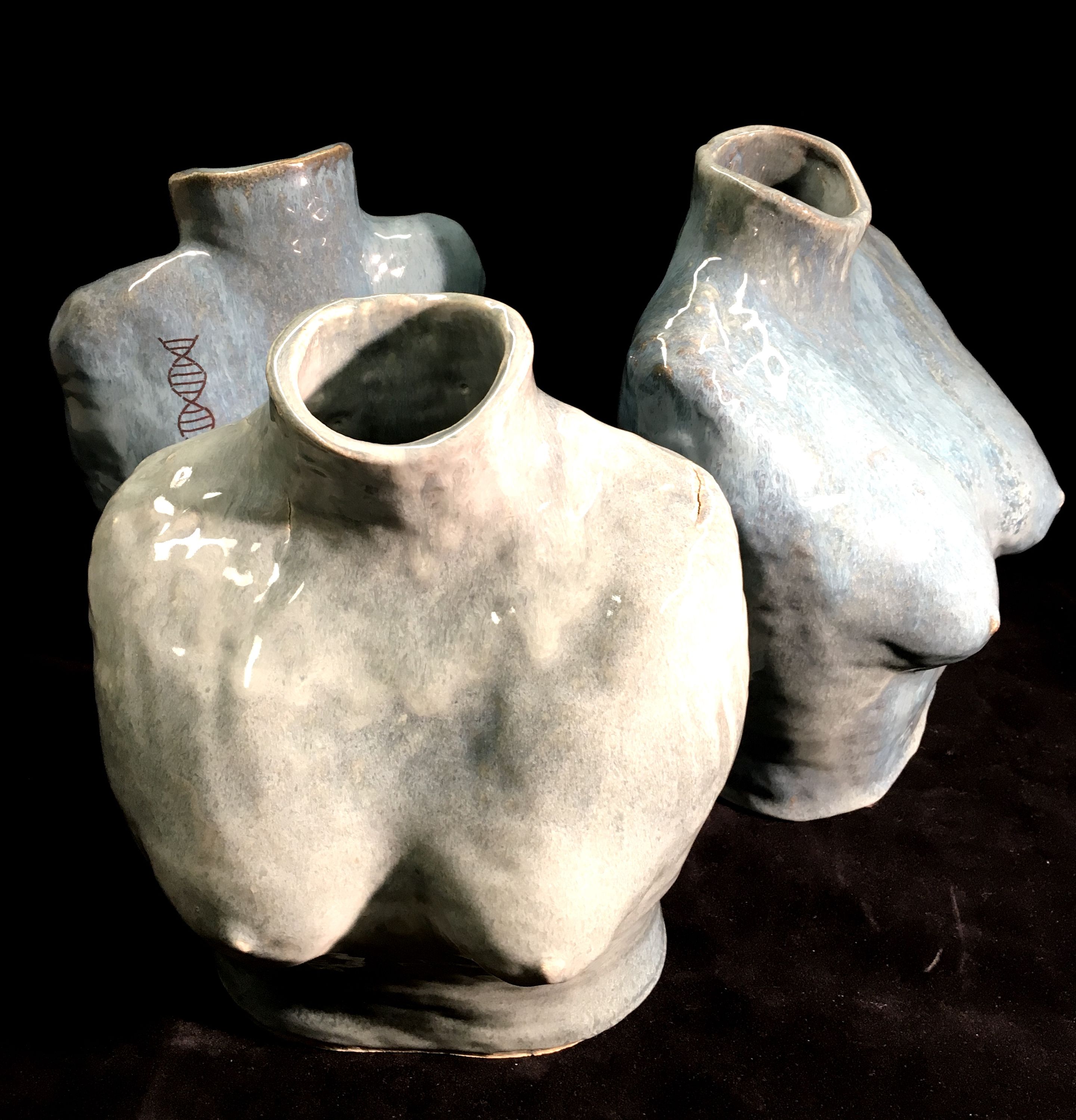 Three ceramic vessels in the shape of a nude woman’s armless torso from the neck to just above the belly button. Their backs are to each other and appear to be duplicates of each other. The vessel closest to the viewer is glazed in a mottled cream-gray while the other two pieces are blue-gray. On the back left shoulder of the piece in the back is a red double helix.