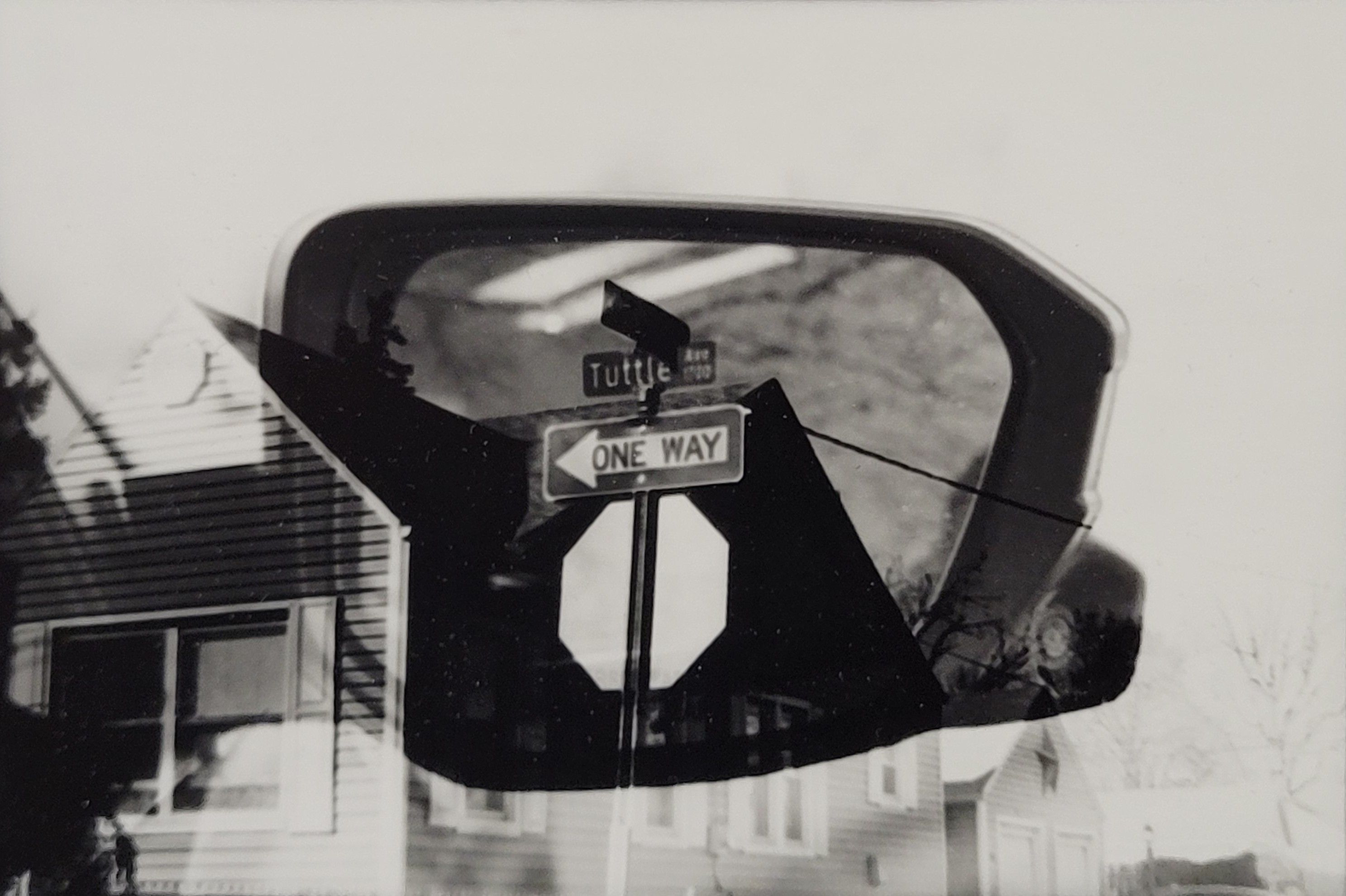 A black and white photograph showing multiple layers of imagery on top of itself. One layer is a street corner, with a one way sign above the back of a stop sign in the center of the photograph. A house is behind the sign, fading into white. Another layer is a side view mirror from a car, this is surrounding the one way sign and making it appear darker than the rest of the image.
