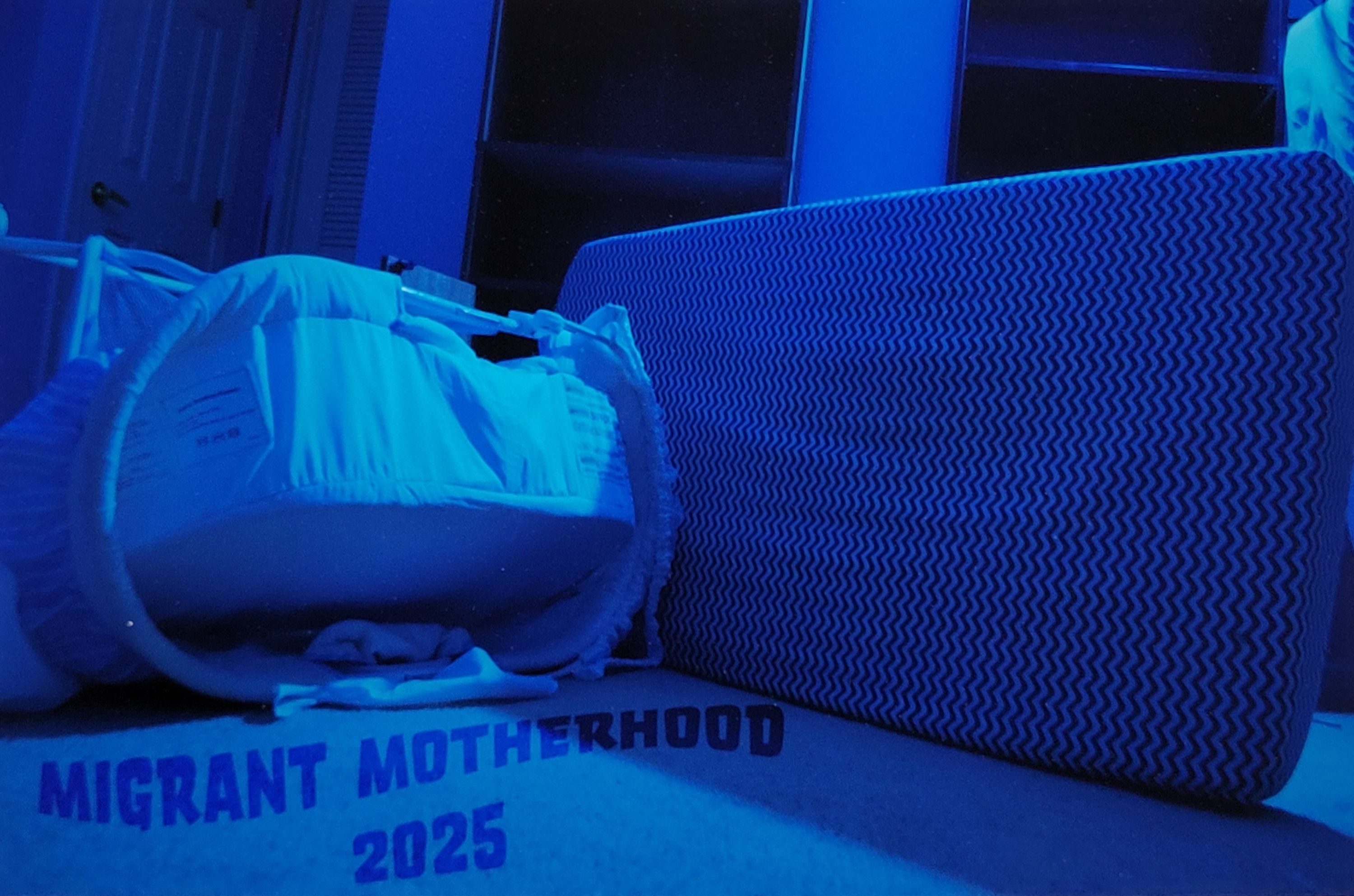 A digital print. A blue tinted photograph depicting a turned over baby bassinet and a toddler bed in a room. The words "Migrant Motherhood 2025" are in text underneath the turned over bedding.