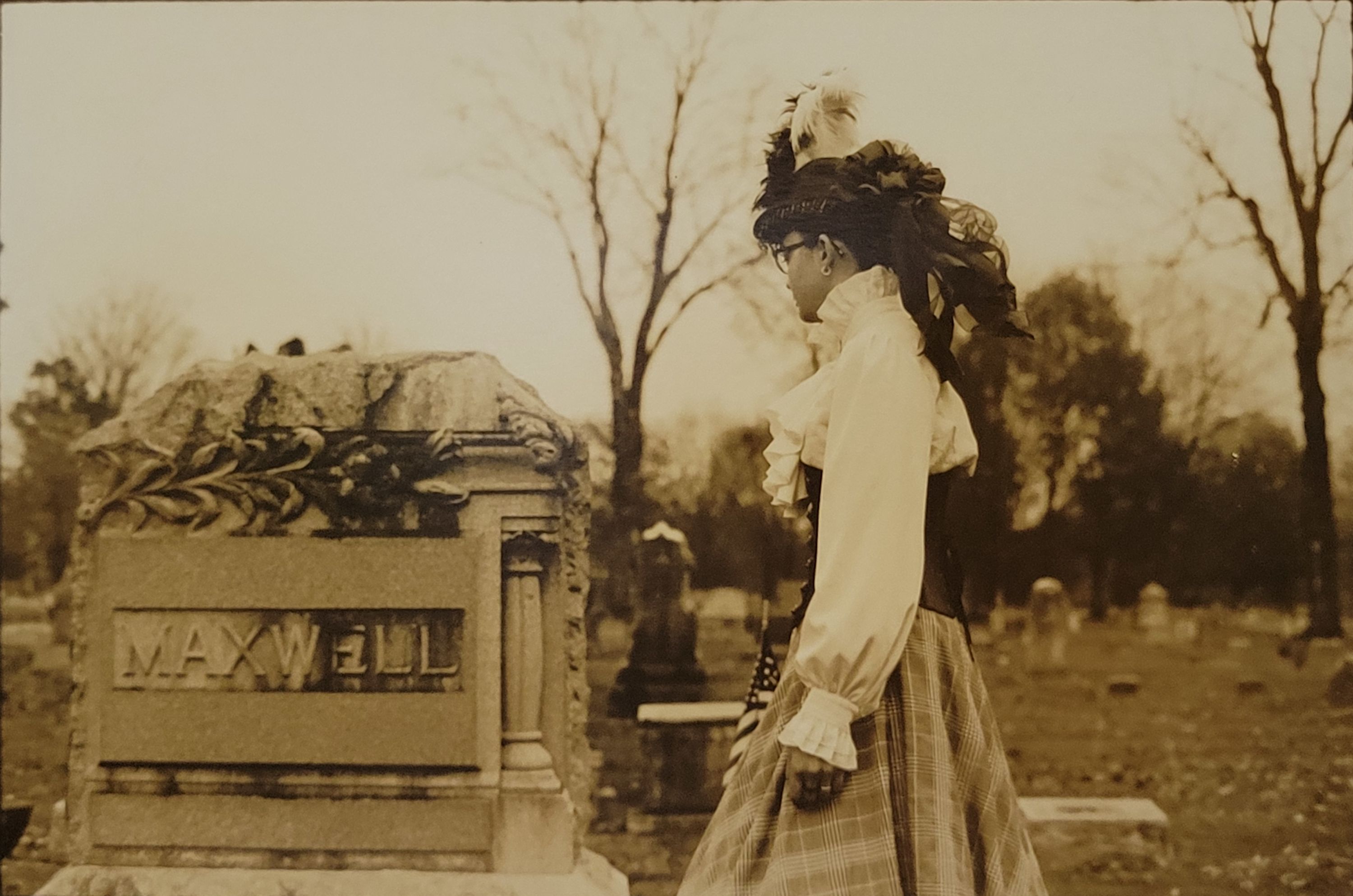 A sepia toned photograph of a young woman in early 1900's attire. She is standing perpendicular to the viewer, with her face turned towards a gravestone on the left side of the photo. The gravestone reads "Maxwell" and is decorated with carved floral and architectural elements. Behind the young woman lies the rest of the cemetery and tall trees, making it feel like time has stopped.